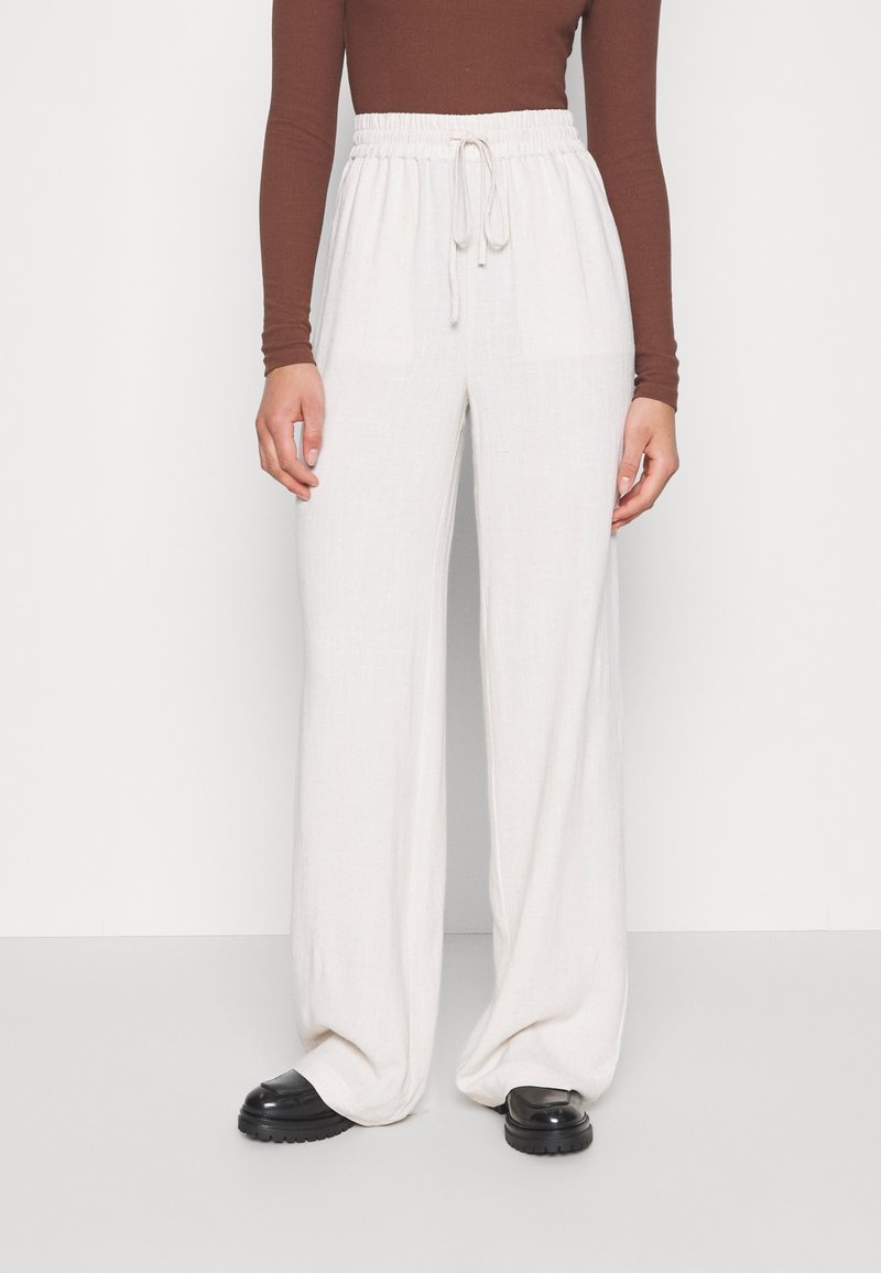 Person som bär lösa vita byxor med dragsko, brun långärmad topp och svarta klumpiga skor, stående mot en enkel bakgrund.