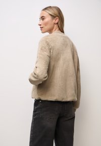 Beige fusk andjursjacka med ribbade hals- och ärmslut, med en avslappnad passform och mjuk textur, parat med mörkgrå jeans.