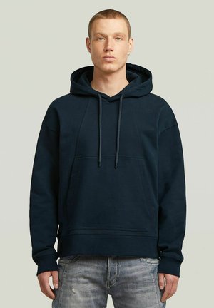 Mann mit kurzen Haaren, der einen dunkelblauen Kapuzenpullover und hellgraue Jeans trägt, steht vor einem schlichten hellen Hintergrund.