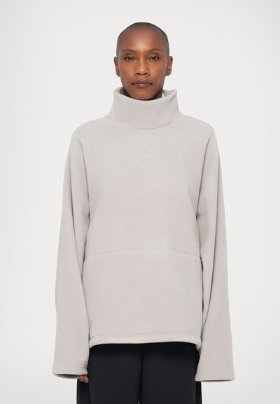 Personne avec une tête rasée portant un pull à col montant en polaire gris clair et un pantalon noir, faisant face à l'avant devant un fond uni.