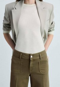 Blazer gris clair à manches retroussées sur un haut crème, associé à un pantalon vert olive avec poches avant et fermeture à bouton sur le devant.