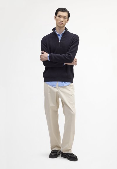 Navy pullover with a half-zip collar, worn over a light blue shirt, paired with cream trousers and black loafers. Smooth texture, relaxed fit.