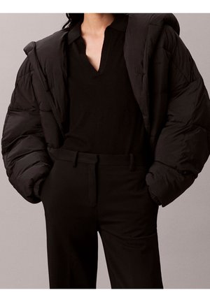 Frau trägt schwarze Steppjacke über schwarzem V-Ausschnitt-Top und hoch taillierte schwarze Hose, mit den Händen in den Taschen vor grauem Hintergrund.