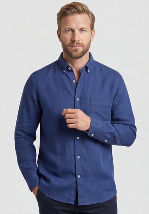 Blonde man met baard, gekleed in een blauw, dichtgeknoopt overhemd met lange mouwen, met één hand op de borst, staand voor een effen lichte achtergrond.