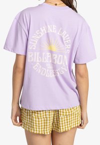 Kvinna i en lavendelfärgad T-shirt med en gul solgrafik och texten "Sunshine Lovers Billabong", kombinerad med gula rutiga shorts.
