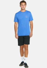 Camisa azul de manga corta con cuello redondo y pequeño logo, combinada con pantalones cortos negros, calcetines blancos y zapatillas deportivas negras.