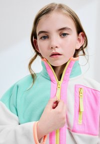 Veste en polaire avec un design à demi-fermeture éclair ; présente des couleurs vert menthe, blanche et rose ; comprend des fermetures éclair jaunes et une poche avant.