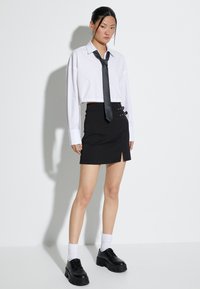Camicia bianca tagliata con cravatta nera, abbinata a una gonna nera con bottoni sul lato e uno spacco, indossata con scarpe nere chunky.