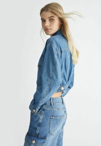 Giubbotto di jeans con maniche lunghe e un taglio corto, dotato di chiusura a bottoni e tasche. Colore azzurro chiaro con una texture morbida.
