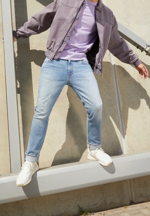 Osoba w jasnoniebieskich dżinsach, białych trampkach, fioletowej koszuli i szarej kurtce balansująca na szarym metalowym belce na zewnątrz.