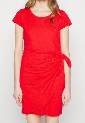 Vrouw die een felrode jurk met korte mouwen draagt, met een detail van een knoop aan de zijkant bij de taille, staand tegen een effen lichte achtergrond.