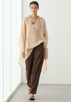 Femme marchant à l'intérieur, vêtue d'un chemisier beige ample à manches larges, d'un pantalon marron foncé, de sandales noires et d'un collier de perles en bois.
