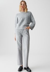 Hellgrauer Strickpullover mit einer strukturierten, flauschigen Oberfläche und elastischem Saum. Kombiniert mit hellgrauen, geraden Jeans und schwarzen Mokassins.