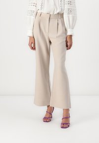 Beige tailored trousers with pleats, flared hem, and smooth texture, paired with purple strappy high-heeled sandals.
