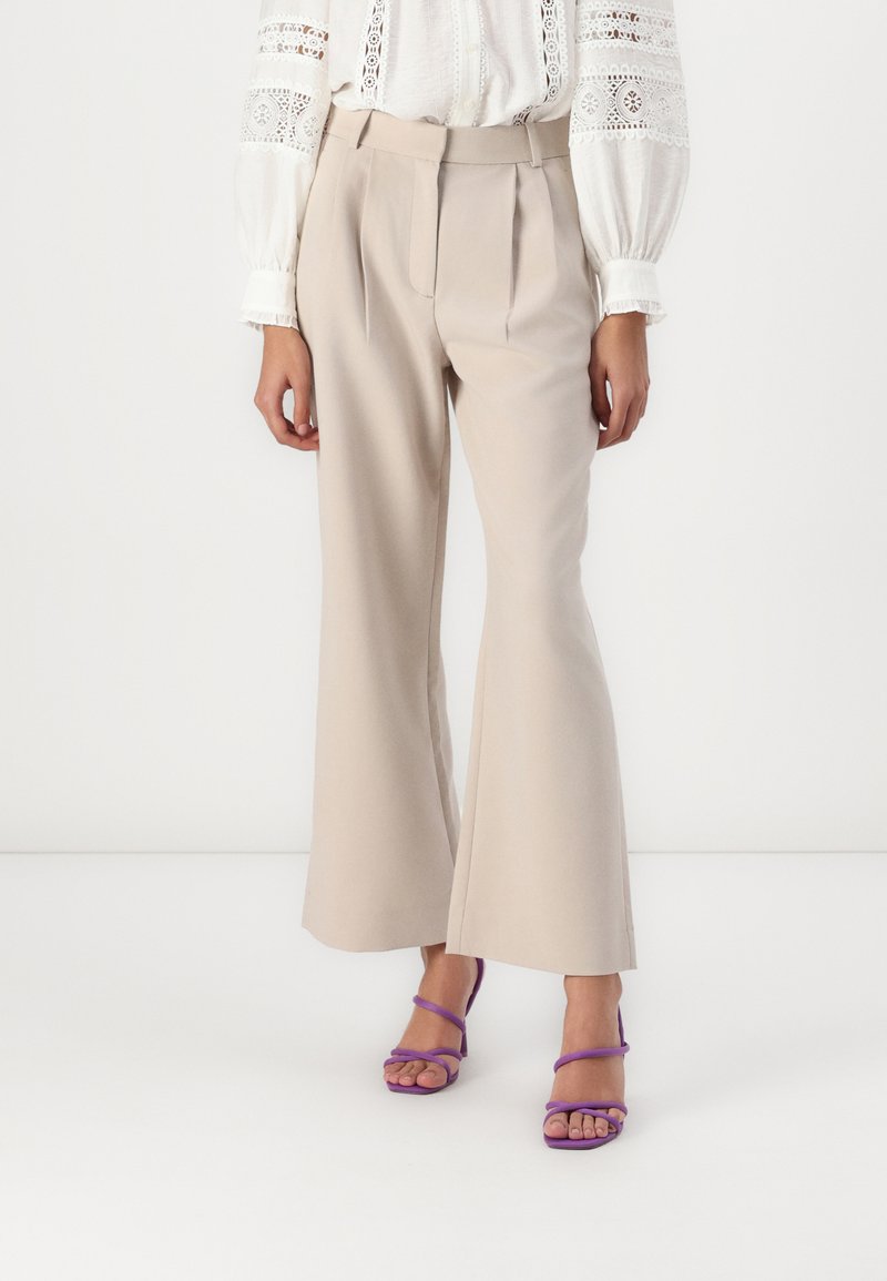 Beige tailored trousers with pleats, flared hem, and smooth texture, paired with purple strappy high-heeled sandals.