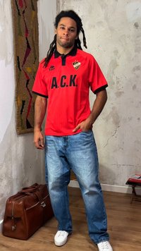 Homme aux dreadlocks portant un maillot de sport rouge A.C.K., un jean bleu et des baskets blanches, debout près d'un sac en cuir marron et d'une tenture murale texturée.