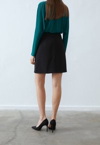 Černá sukně s přiléhavým střihem, kombinovaná s topem se dlouhým rukávem v barvě smaragdové. Celkový vzhled doplňují černé špičaté vysoké podpatky. Neutrální pozadí.