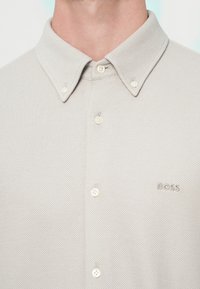 Világosszürke galléros ing texturált anyagból, finom mintával, öt gombbal és egy kis hímzett logóval a mellrészen.