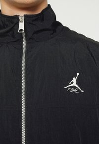 Veste coupe-vent noire avec col montant, fermeture éclair argentée et logo Jumpman brodé avec le texte "Flight" sur la poitrine. Texture froissée.