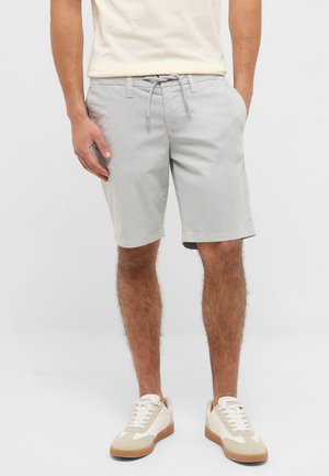 Mann trägt hellgraue Shorts mit Kordelzug und weiße Turnschuhe, steht mit den Händen in den Taschen vor einem einfarbigen Hintergrund.