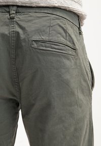 Zbliżenie na oliwkowe spodnie z kieszenią z tyłu i szlufkami na pasek, noszone z lekkim, dzianinowym swetrem.