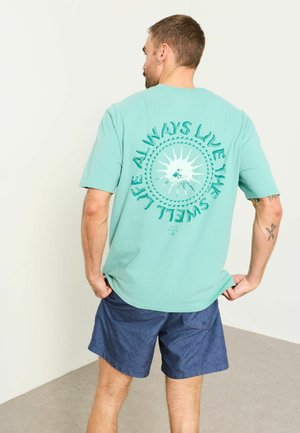 Mand med kort hår iført en turkis T-shirt med sol og teksten "Live in the swell life always" samt blå mønstrede shorts.