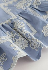 Világoskék szövet fehér hímzett virágmintákkal és díszített szegélyekkel, redőzött, puha és sima tapintású.