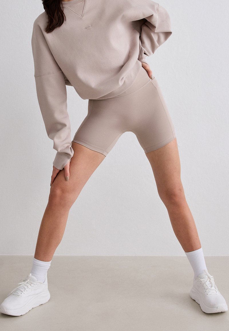 Mujer con sudadera beige y shorts de ciclista a juego, calcetines blancos y zapatillas blancas, de pie con las piernas separadas y una mano en la cadera.
