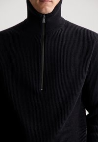 Maglione nero a costine con colletto alto e zip a un quarto, realizzato in un tessuto testurizzato che crea un aspetto morbido e accogliente.