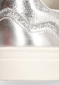 Zapatilla metálica plateada con un acento ondulado brillante a lo largo del borde superior, con un acabado texturizado y una suela de goma blanca.