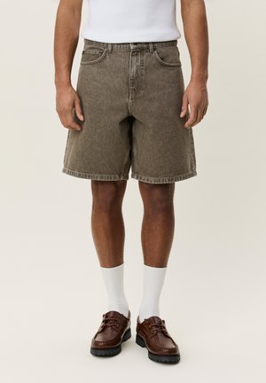 Man draagt bruine denimshorts, witte sportsokken, bruine leren schoenen en een wit mouwloos shirt, staand tegen een effen achtergrond.
