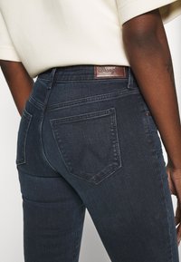 Mörka denimjeans med en figursydd design, som har en brun läderetikett vid midjan och två bakfickor med en distinkt form.
