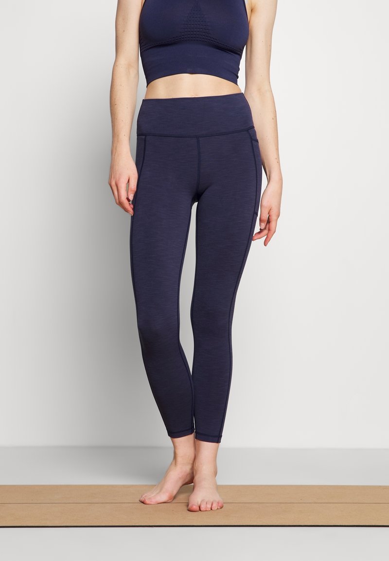 Kvinna står barfota på en yogamatta iförd mörkblå högmidjade leggings och en matchande kort atletisk topp.