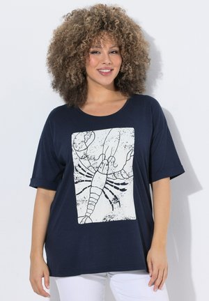 Frau mit lockigem Haar lächelt, trägt ein marineblaues T-Shirt mit großem weißem Hummer-Design, kombiniert mit weißen Hosen.