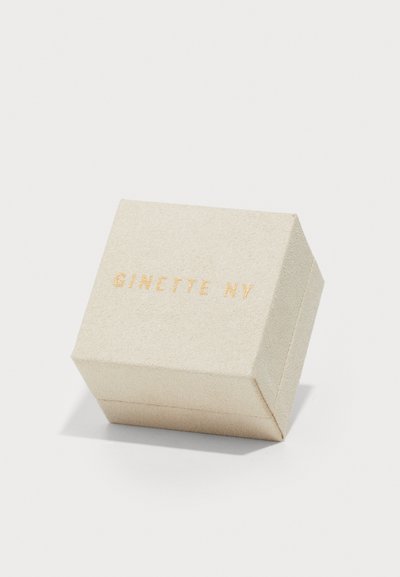 Ruutjalne ehtelaegeri pehme kreemjas materjalis, millel on peen tekstuur, kuldne embosseeritud "GINETTE NY" peal ja puhas, minimalistlik disain.