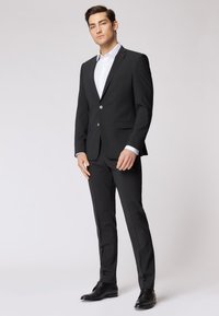 Abito nero con un design slim-fit, caratterizzato da un bottone singolo, rever a punta e una camicia bianca sotto. Scarpe eleganti nere.
