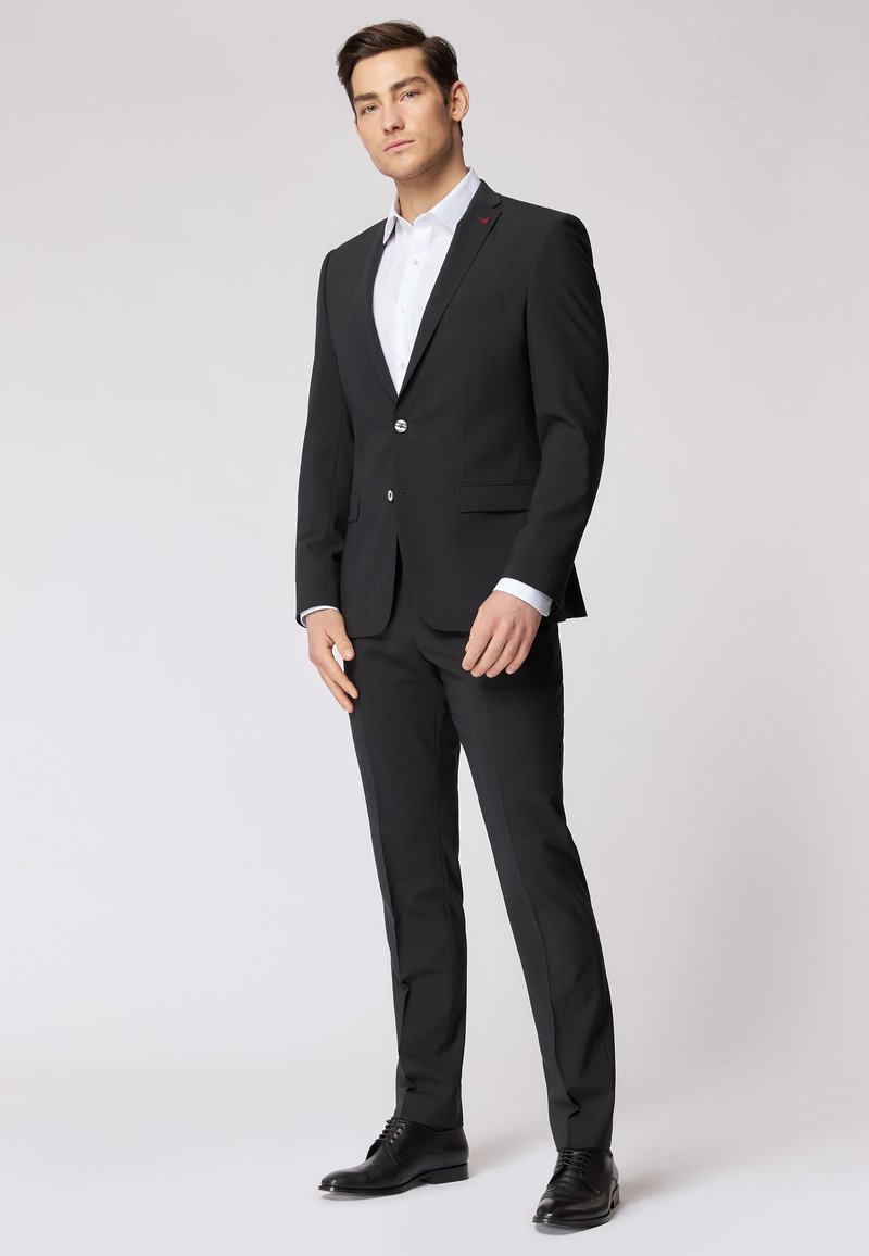 Abito nero con un design slim-fit, caratterizzato da un bottone singolo, rever a punta e una camicia bianca sotto. Scarpe eleganti nere.