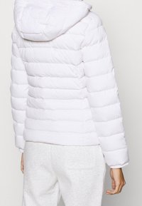 Biały puchowy płaszcz z poziomym przeszyciem, z doczepionym kapturem i ściągaczami na rękawach. Miękka, wypełniona tekstura; noszony z jasnoszarymi spodniami dresowymi.