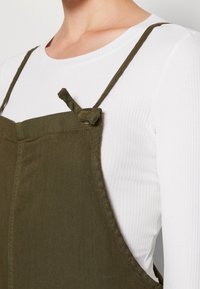 Detailní záběr na osobu v bílé žebrované halence s dlouhým rukávem pod olivově zeleným overalovým šaty s uzlem na rameni.