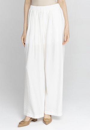 Kobieta nosząca luźne, białe spodnie o szerokich nogawkach z elastycznym pasem i beżowe, szpiczaste szpilki ze złotymi klamrami.