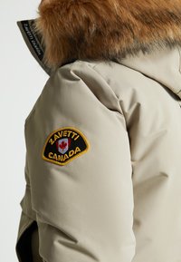 Bēšā pūkainā jaka ar kažokādas oderi kapuces iekšpusē. Iespējo melno un zeltīgo izšuvumu uzlauku ar uzrakstu "ZAVETTI CANADA" un kļavas lapas dizainu.