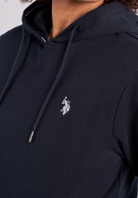 Mörkblå hoodie med mjuk textur, med en vit broderad logotyp på bröstet och silverfärgade ändar på dragsstrings.