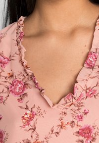 Blusa floreale rosa con scollatura a V e bordo arricciato. Realizzata in tessuto leggero con motivi floreali rosa e marroni.
