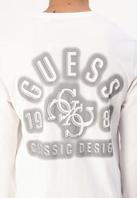 Bijela majica dugih rukava siva grafika s natpisom "GUESS", godinom "1981" i kružnim akcentima u hrabrom, modernom fontu.