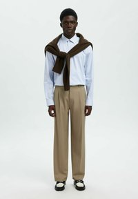 Camisa a rayas azul y blanca, suéter marrón atado al cuello, pantalones beige, zapatos en blanco y negro. Diseño minimalista, corte ajustado.