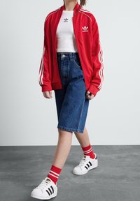 Czerwona sportowa kurtka z poliestru z białymi paskami, biały T-shirt, szorty jeansowe z wysokim stanem, czerwone skarpetki z białymi paskami oraz białe sneakersy z czarnymi paskami.