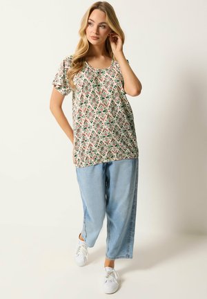 Frau trägt ein kurzärmliges Oberteil mit Blumenmuster, hellblaue Jeans mit weitem Bein und weiße Turnschuhe, steht mit einer Hand nahe am Gesicht.