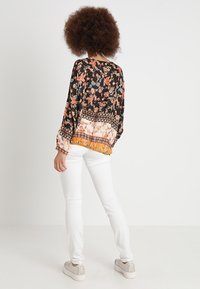 Femme aux cheveux bouclés vue de dos, portant un chemisier à motifs floraux, un pantalon blanc et des baskets beige sur un fond uni.