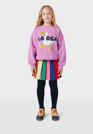 Ružový sveter s grafickým motívom kačky a textom "LA OCA"; viacfarebná plisovaná sukňa; čierne pančuchy; farebné topánky s žltými akcentmi.