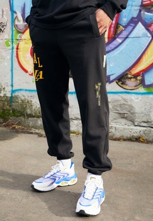 Pantaloni della tuta neri con stampa grafica gialla, tasche laterali e dettagli. Abbinati a scarpe da ginnastica bianche e blu. Tessuto texturizzato.